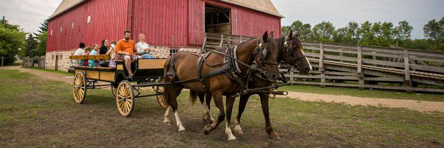 Un attelage de chevaux tire un chariot avec des visiteurs près d’une grange rouge.