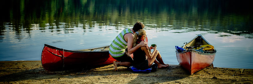 A couple is sitting by the lake near canoes.