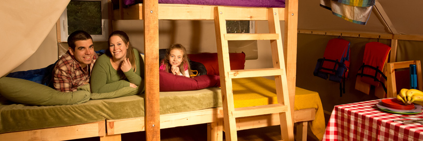 Parents étendus dans le bas du lit à deux étages de la tente oTENTik, pendant que leurs deux enfants s’amusent dans le plus petit lit du deuxième étage.