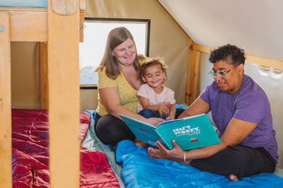A couple with their child read a book together. They are sitting on an oTENTik bunkbed covered with sleeping bags.