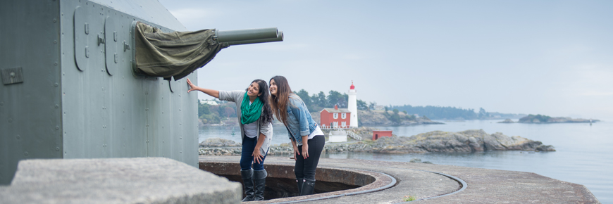 Deux jeunes femmes regardent le canon bitube de 6 livres. Au loin, le phare de Fisgard.