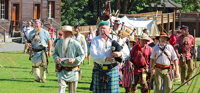 Group of historic costumed volunteers marching, led by a bagpipe player.