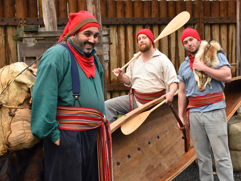 Three costumed interpreters wearing Voyageur sashes, capots, and holding wooden paddles.