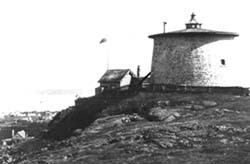Une photo en noir et blanc de la Tour-Martello-de-Carleton et la maisonnette. Le port et des maisons sont à l'arrière-plan.