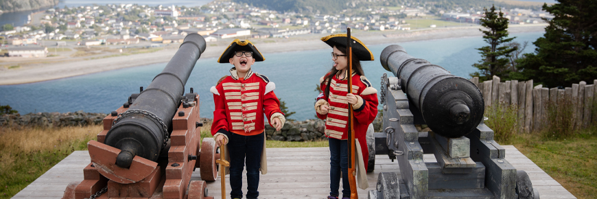 two children in historic military costume laughing next to two cannons overlooking a coastal town