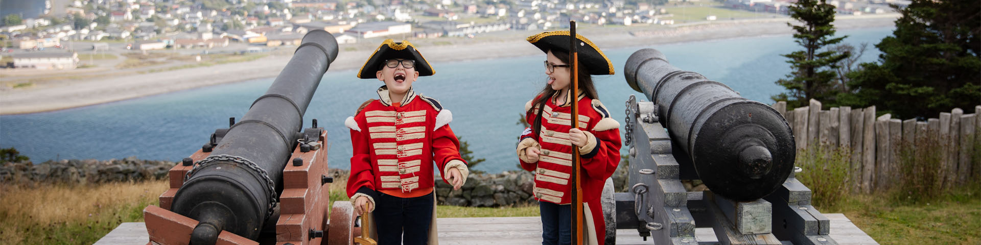 deux enfants en costume militaire historique riant à côté de deux canons surplombant une ville côtière