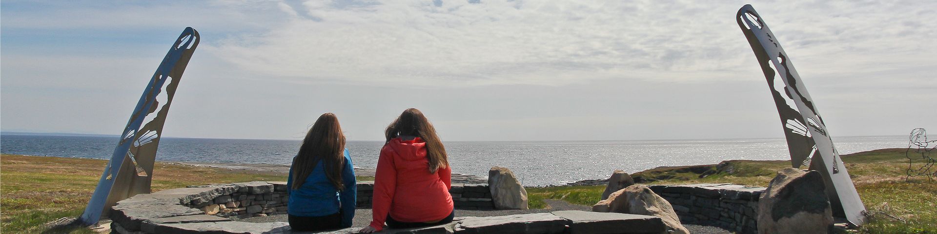 2 people sit on a circular stone bench and look toward the coast at Port au Choix National Historic Site