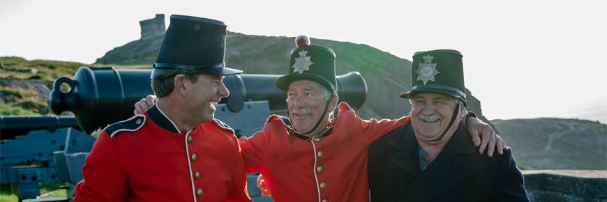 Trois hommes en costume historique souriant avec un canon et la tour Cabot à l'arrière-plan