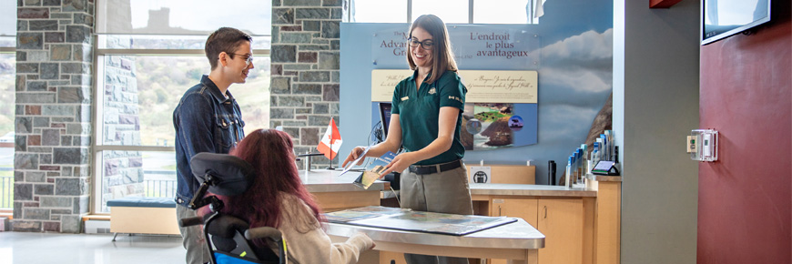a Parks Canada employee shows two people, one in a wheelchair and the other standing, a brochure from behind a desk