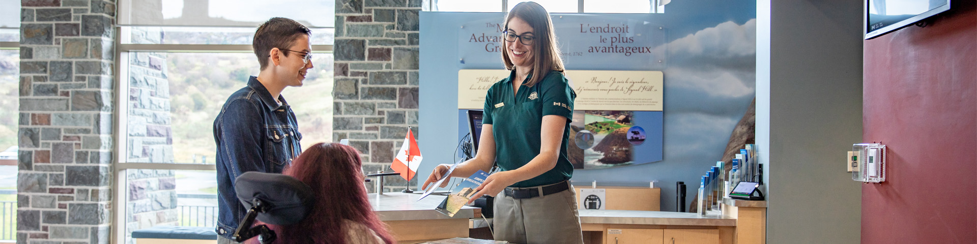 a Parks Canada employee shows two people, one in a wheelchair and the other standing, a brochure from behind a desk