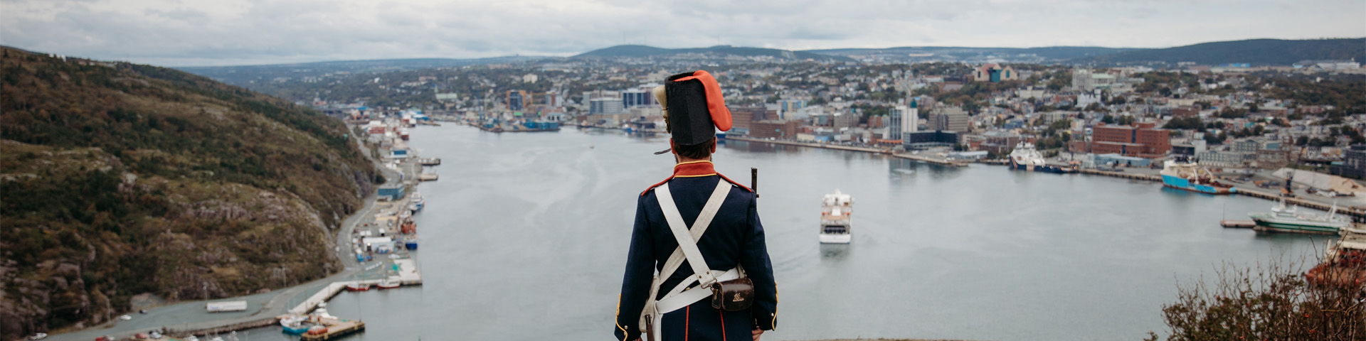 a man in an historic military uniform, overlooking a city harbour.