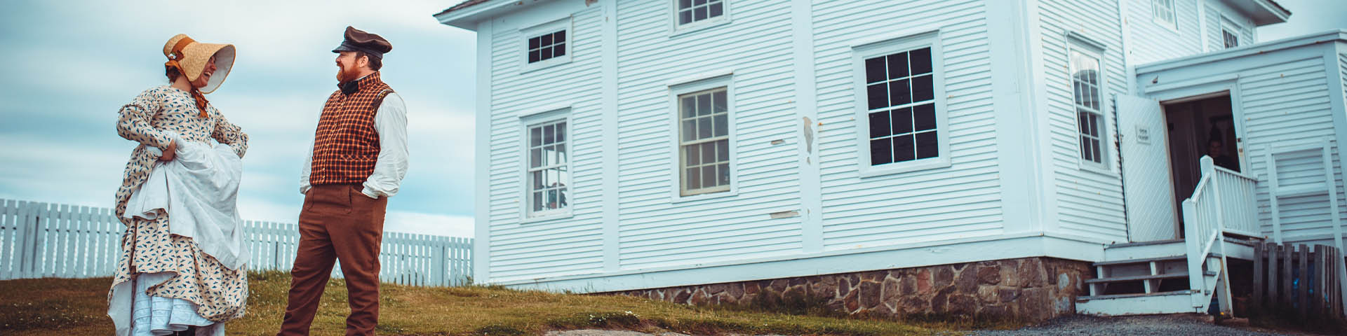 deux individus en costume historique, riant à l'extérieur d'une maison blanche en bois.