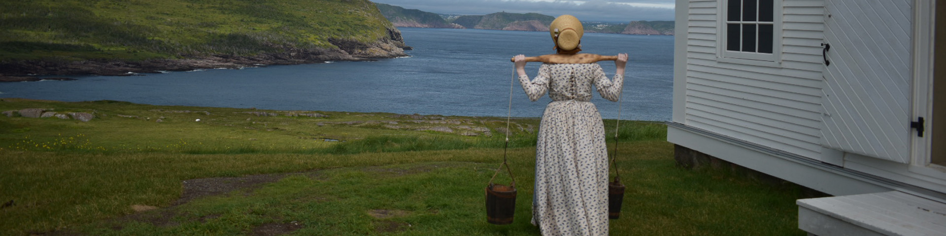 une femme en costume historique portant de l'eau regarde une vue de la côte près d'un phare