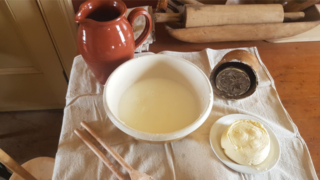 supplies for making butter on a table, including spoons, a bowl, a stamp, and a ceramic jug
