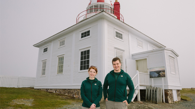 two Parks Canada employees standing outside of a white and red, wooden house
