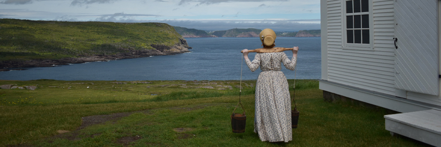 une femme en costume historique portant de l'eau regarde une vue de la côte près d'un phare