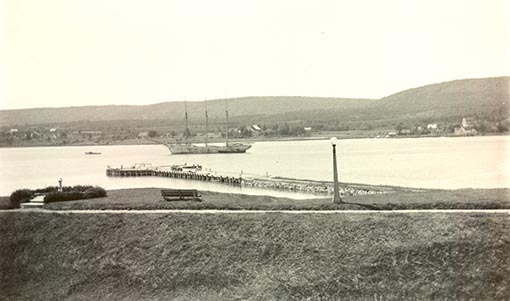 An historic photo of the Queen’s wharf in the Annapolis River, with a tall ship in the background.