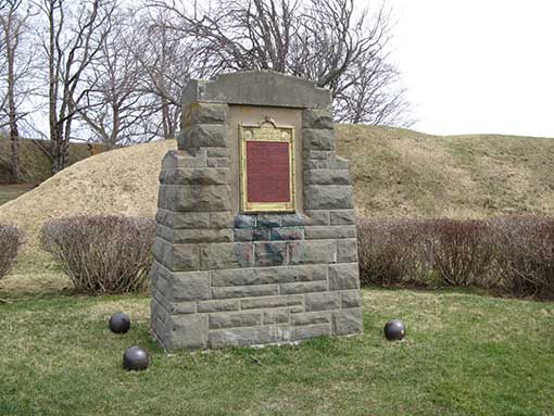 Monument en pierre avec une plaque marron. Quatre boulets de canon se trouvent dans l’herbe aux quatre coins de sa base.