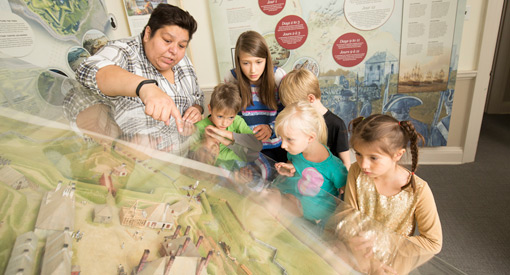  A woman and a group of children look at a model of the fort that is under plexiglass.