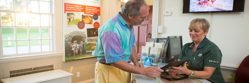 Au comptoir du Centre d’accueil au lieu historique national du Fort-Anne, un membre du personnel de Parcs Canada montre quelque chose sur une tablette à un visiteur. 