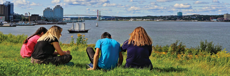 A small group sitting on the grass at Georges Island watching a tall ship in Halifax with Halifax, Purdy’s wharf, Macdonald bridge, and Dartmouth in the background. 