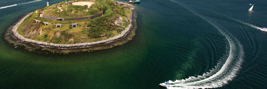 An aerial view of Georges Island showing the ocean facing side of the island and a speedboat circling the island in the harbour. 