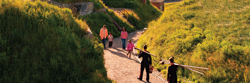 Une jeune famille rencontre des guides-interprètes costumés transportant de longs bâtons et des chaudières de poudre à canon le long d’un sentier au fort Charlotte.