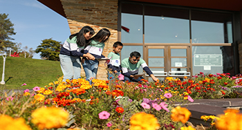 A family of four kneels next to a flower bed in front of a stone building with windows