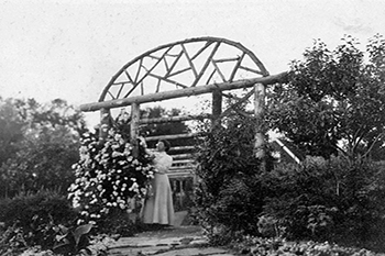 A woman stands on a cobble path under a wooden arch in a garden reaching upwards touching flowers