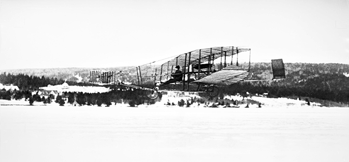 A side view of the Silver Dart flying over a frozen lake