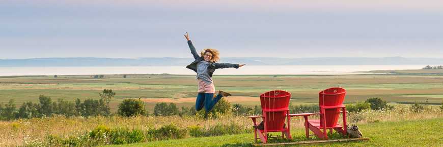 A woman jumping in the air near the red chairs with the Landscape of Grand-Pré in the background