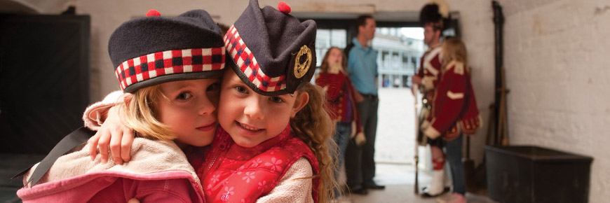 Two children wear soldier’s hats and smile for the camera while their parents and older sibling talk with an interpreter in 78th Highlander uniform in the background at Halifax Citadel.