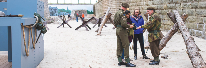 Two female visitors and two soldiers stand discussing a gas mask in foreground on the sand and among the barricades and a boat replica from the “Storm the Beach D-Day Exhibit” as other visitors explore the exhibit in the background.