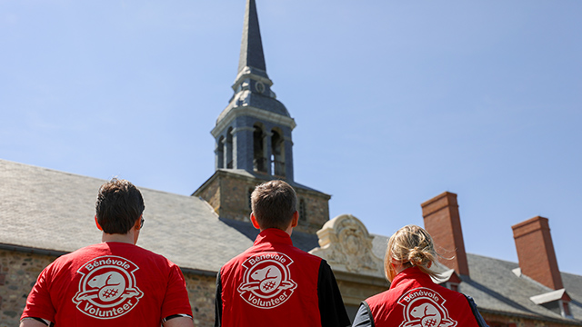 Three people look up at a clock tower, they are wearing red volunteer vests with a Parks Canada logo.