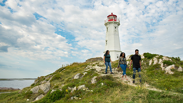 Three people walk on a hiking trail with an historical lighthouse in the background on a sunny day