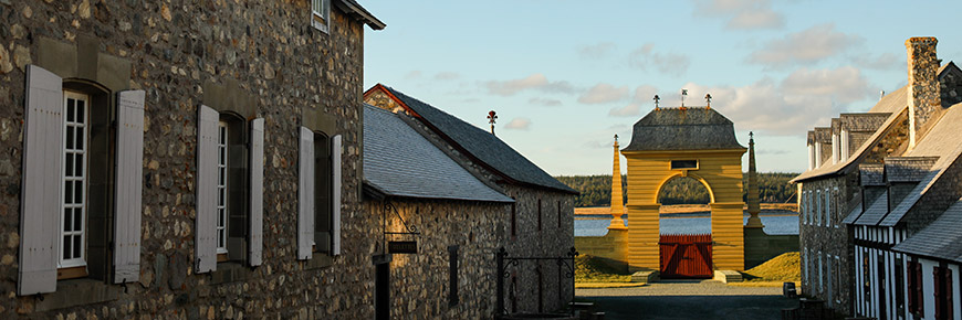 A street view at the Fortress of Louisbourg with 18th-century buildings. At the end of the street is the yellow Frédéric gate and the Louisbourg Harbour. 