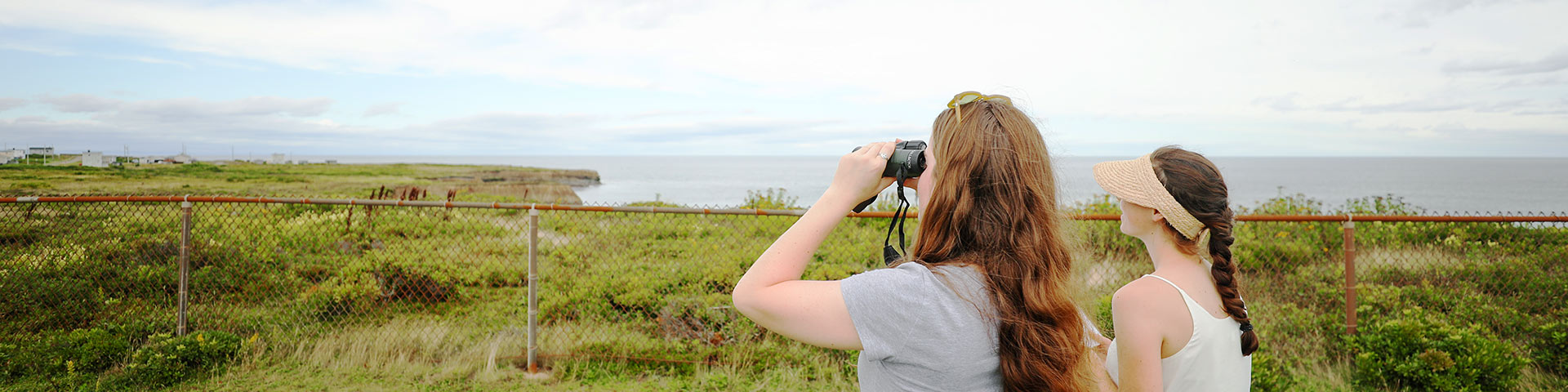 Two women stand by a cliff on the ocean, one looks through binoculars