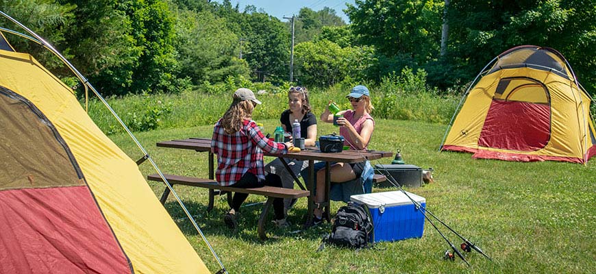 Three young women sit at a picnic table between two tents on their campsite with a cooler and fishing poles visible beside them