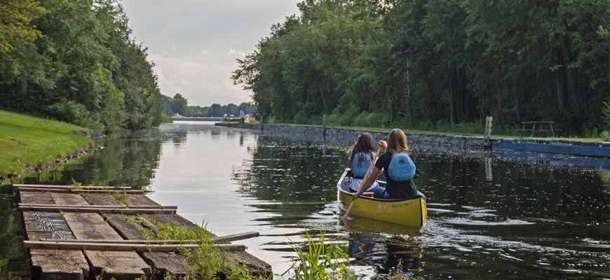 Two women in a canoe paddling away from the dock on a narrow canal heading towards a lock on the horizon