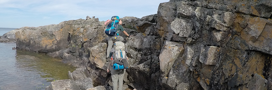 Deux randonneurs escaladent une petite paroi rocheuse le long du lac Supérieur, tout en portant des sacs à dos.