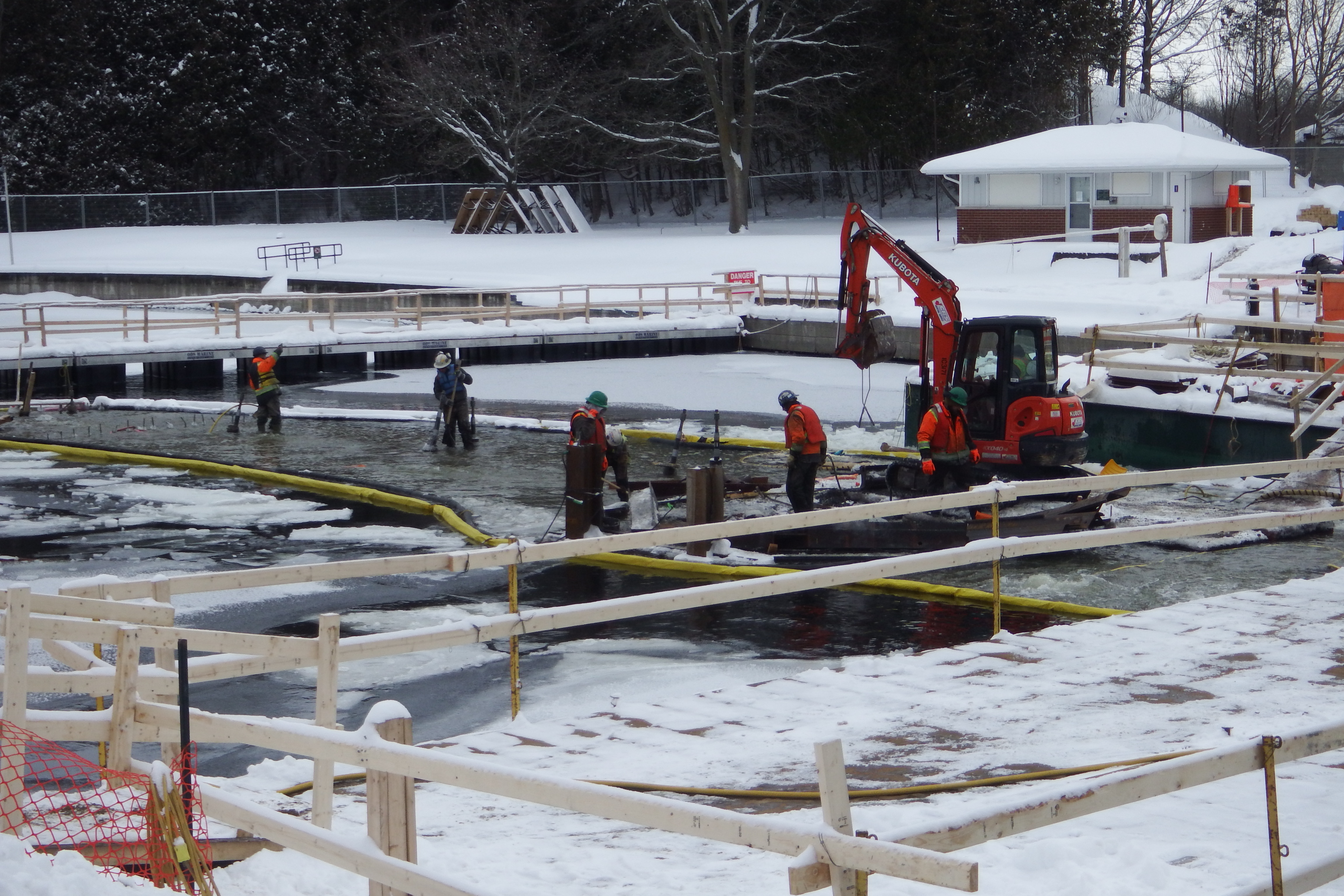 A snowy construction site. Workers in the water with a mini excavator nearby.