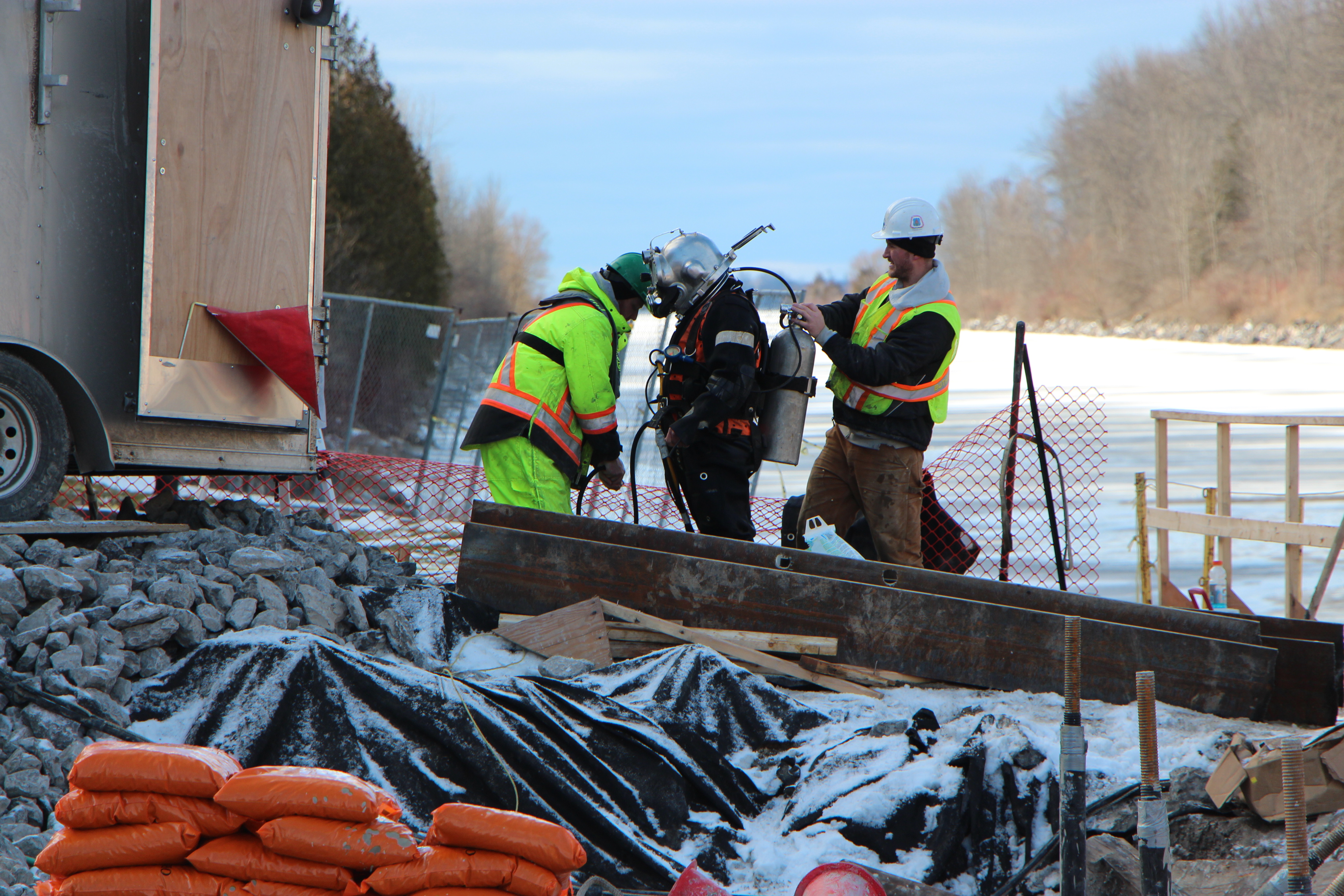 A snowy construction site. Two people help to check the equipment of a hardhat diver.