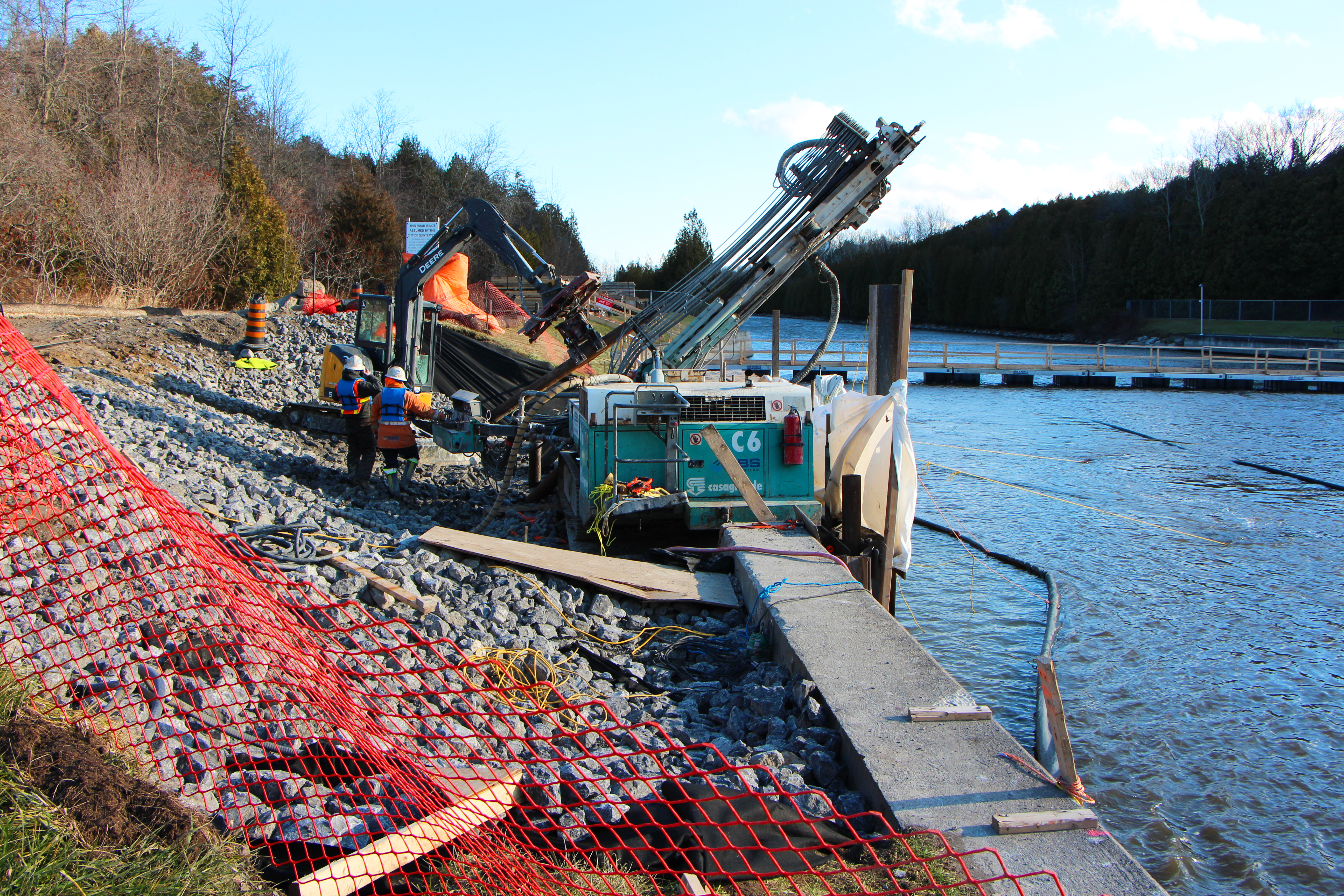 A drill rig sits adjacent to the shoreline drilling diagonal holes into the rock below to accept new micropiles.