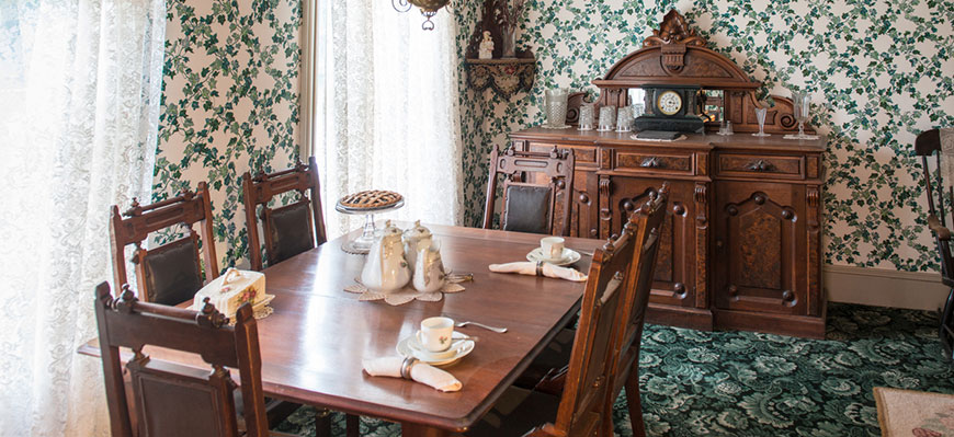 Antique dinning room set in room with green printed carpet and green pattern wallpaper. 