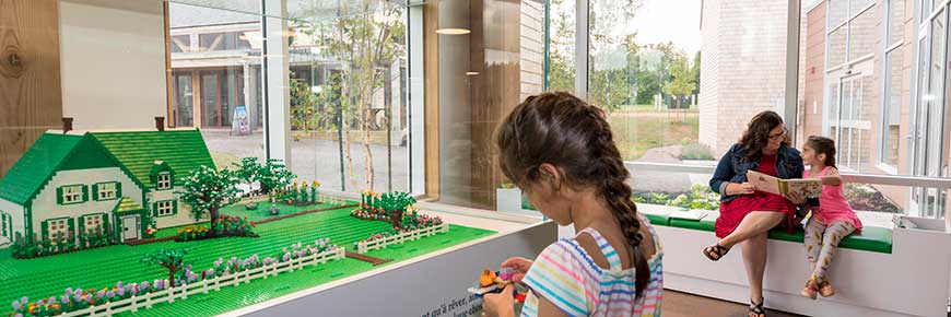 A young girl plays at the block model exhibit at Green Gables Visitor Centre, while a woman and young girl sit on a bench reading a book in the background. 