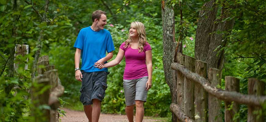 A young male wearing a blue shirt walks with a young female wearing a purple shirt on the pathway of Lover's Lane, Green Gables Heritage Place, surrounded by green trees. 