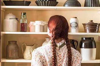 Une imitatrice d'Anne Shirley avec deux tresses rouges se tient dos à la caméra devant une armoire ouverte remplie d'ustensiles de cuisine au site patrimonial Green Gables.