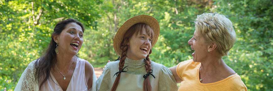 An Anne impersonator with red braids and a sun hat stands in the forest laughing with two female visitors at Green Gables Heritage Place. 