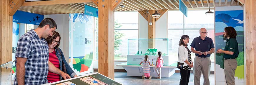 Dans une vue grand angle de l'espace ouvert et lumineux du centre d'accueil de Green Gables, un couple à gauche lit un panneau, deux jeunes filles jouent devant une maquette de bloc et un autre couple discute avec un guide de Parcs Canada en uniforme. 