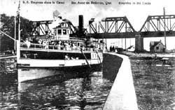 Le bateau S.S. Empress avec plusieurs passagers sur le pont sortant du canal de Sainte-Anne-de-Bellevue vers l'amont. À l'arrière-plan, le pont de la voie ferroviaire.